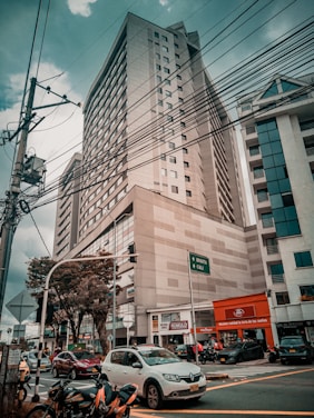 A busy urban street scene with a tall office building in the background. The sky is overcast, and power lines crisscross the upper part of the image. Several cars and motorcycles are at the foreground intersection, and there are street signs pointing towards Bogota and Cali. Trees are present along the sidewalk, and various small shops are visible at street level.