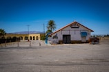 An old, worn-down motel with a tiled roof is situated beside a parking lot. The facade appears weathered, with a closed entrance and a small blue awning. A palm tree stands nearby, and in the background, there are mountains and a clear blue sky. An old dumpster with graffiti is positioned on the right side.