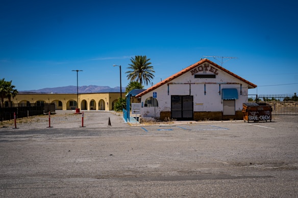 An old, worn-down motel with a tiled roof is situated beside a parking lot. The facade appears weathered, with a closed entrance and a small blue awning. A palm tree stands nearby, and in the background, there are mountains and a clear blue sky. An old dumpster with graffiti is positioned on the right side.