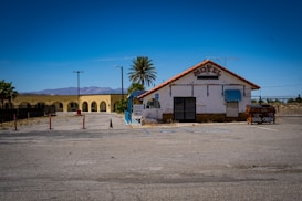 An old, worn-down motel with a tiled roof is situated beside a parking lot. The facade appears weathered, with a closed entrance and a small blue awning. A palm tree stands nearby, and in the background, there are mountains and a clear blue sky. An old dumpster with graffiti is positioned on the right side.
