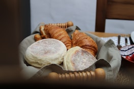 A basket filled with freshly baked pastries, including croissants and English muffins, placed on a wooden table. The basket is lined with a light gray cloth, and other items such as a bowl with a pattern and a plate can be seen in the background.