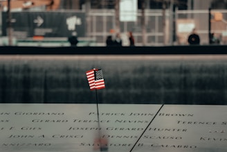 An engraved memorial surface with a small American flag placed in one of the etched names. The background shows a blurred view of people and a construction area.