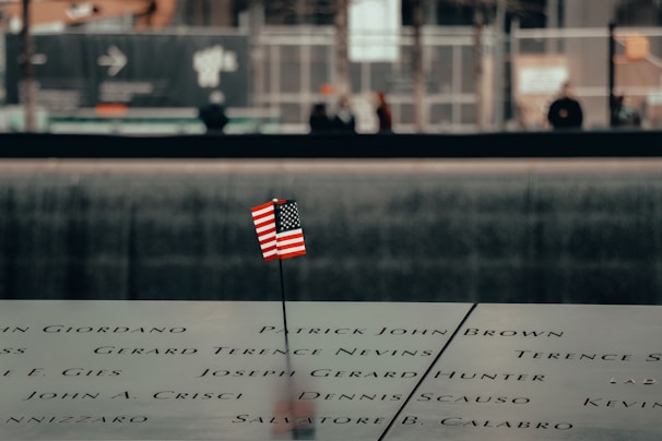 An engraved memorial surface with a small American flag placed in one of the etched names. The background shows a blurred view of people and a construction area.