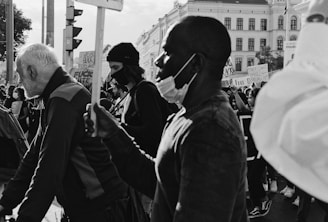 A group of people is participating in a protest, holding signs and wearing face masks. The crowd appears diverse and engaged in the event taking place in an urban setting with buildings in the background.