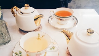 A table set for tea with two ornate teapots and matching teacups. The teapots and cups are white with gold accents. The teacups contain warm beverages, one light yellow and another darker amber.