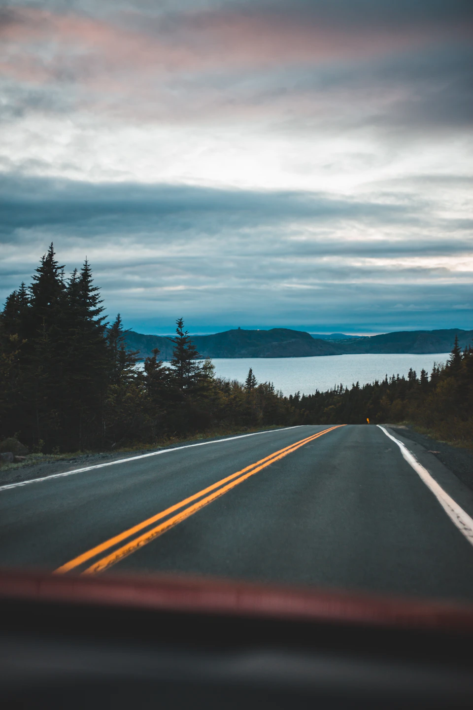 The image shows a winding road with two lanes, one on each side, that stretches into the distance and disappears over a body of water. The sky above is filled with clouds, casting a soft light over the scene. On either side of the road are trees, their green foliage contrasting against the cloudy sky. In the background, mountains rise up to meet the horizon line, adding depth and scale to the image.