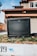 An old television set is placed outside on a small patch of grass in a residential area. The background features a suburban house with a gray facade and brown trimming. The image is slightly blurred, creating a nostalgic or vintage feel.