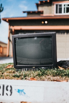 An old television set is placed outside on a small patch of grass in a residential area. The background features a suburban house with a gray facade and brown trimming. The image is slightly blurred, creating a nostalgic or vintage feel.