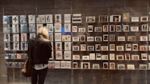A person with short blonde hair, wearing a leather jacket, stands in front of a display of various artworks and postcards arranged in a grid on a wall. The pieces include illustrations, photography, and graphic designs, all enclosed within glass casing. An assortment of colorful, abstract, and figurative art is visible.