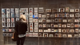 A person with short blonde hair, wearing a leather jacket, stands in front of a display of various artworks and postcards arranged in a grid on a wall. The pieces include illustrations, photography, and graphic designs, all enclosed within glass casing. An assortment of colorful, abstract, and figurative art is visible.