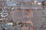Heavily rusted metal surface with peeling paint, varying in shades from dark brown to light grey. The surface is marked with worn-out spots exposing the layers beneath, and graffiti is visible in white.