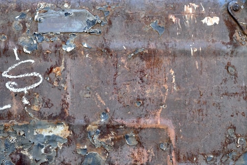 Heavily rusted metal surface with peeling paint, varying in shades from dark brown to light grey. The surface is marked with worn-out spots exposing the layers beneath, and graffiti is visible in white.