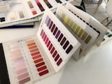 A variety of color swatch books are open on a table, displaying organized grids of color samples. The colors range from light pastels to vibrant reds and purples, as well as shades of green. The swatches are labeled with numbers and arranged systematically across several pages.