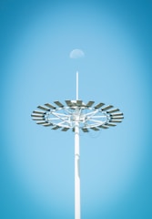 A tall light pole with a circular arrangement of solar panels is seen against a clear blue sky. The moon is faintly visible above the structure.
