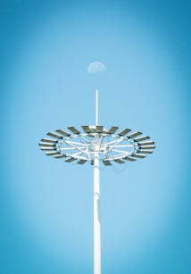 A tall light pole with a circular arrangement of solar panels is seen against a clear blue sky. The moon is faintly visible above the structure.