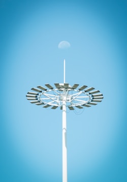 A tall light pole with a circular arrangement of solar panels is seen against a clear blue sky. The moon is faintly visible above the structure.