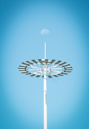 A tall light pole with a circular arrangement of solar panels is seen against a clear blue sky. The moon is faintly visible above the structure.