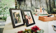 A table is set with framed photographs. One frame shows a couple laughing joyfully, while the other displays an overhead view of a person walking through a forest. In the background, a large image of a couple walking in a wooded area is present. A woven basket sits on the table alongside some pens and flowers.