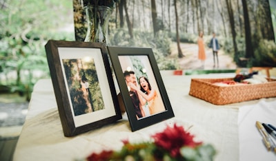 A table is set with framed photographs. One frame shows a couple laughing joyfully, while the other displays an overhead view of a person walking through a forest. In the background, a large image of a couple walking in a wooded area is present. A woven basket sits on the table alongside some pens and flowers.