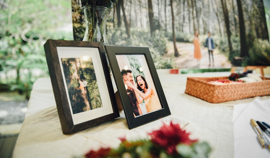 A table is set with framed photographs. One frame shows a couple laughing joyfully, while the other displays an overhead view of a person walking through a forest. In the background, a large image of a couple walking in a wooded area is present. A woven basket sits on the table alongside some pens and flowers.