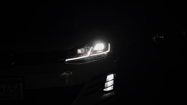 A car headlight is illuminated in the darkness, casting bright light and revealing a small part of the vehicle against a dark background. The image captures the LED detailing and sleek design of the headlight and front grille.