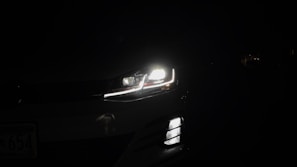 A car headlight is illuminated in the darkness, casting bright light and revealing a small part of the vehicle against a dark background. The image captures the LED detailing and sleek design of the headlight and front grille.