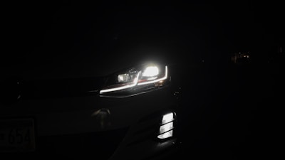 A car headlight is illuminated in the darkness, casting bright light and revealing a small part of the vehicle against a dark background. The image captures the LED detailing and sleek design of the headlight and front grille.