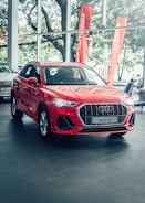 A bright red Audi Q3 is prominently displayed in a car showroom with sleek design elements and visible branding. Large windows allow natural light to illuminate the car, which is positioned on a polished tile floor. The background features green trees and orange promotional banners outside the windows.