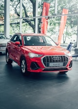 A bright red Audi Q3 is prominently displayed in a car showroom with sleek design elements and visible branding. Large windows allow natural light to illuminate the car, which is positioned on a polished tile floor. The background features green trees and orange promotional banners outside the windows.