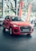 A bright red Audi Q3 is prominently displayed in a car showroom with sleek design elements and visible branding. Large windows allow natural light to illuminate the car, which is positioned on a polished tile floor. The background features green trees and orange promotional banners outside the windows.