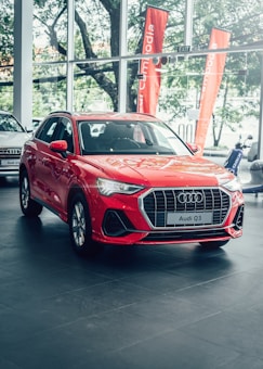 A bright red Audi Q3 is prominently displayed in a car showroom with sleek design elements and visible branding. Large windows allow natural light to illuminate the car, which is positioned on a polished tile floor. The background features green trees and orange promotional banners outside the windows.