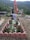 A grave adorned with red and white flowers is surrounded by a low pink brick border. A photograph and a flag are positioned at the head of the grave. The background features a scenic view of lush green hills and a village with scattered houses.