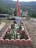 A grave adorned with red and white flowers is surrounded by a low pink brick border. A photograph and a flag are positioned at the head of the grave. The background features a scenic view of lush green hills and a village with scattered houses.