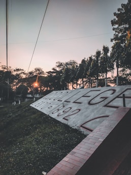 A large sign with the text 'COLLEGE ESTD. 1892' is prominently displayed against a backdrop of trees. The sun is setting, casting a warm glow over the scene. Power lines and foliage are silhouetted against the pastel sky, adding depth to the composition.