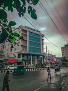 A bustling urban street scene featuring a multi-story building with a glass facade and shop signs. The road in front showcases a few pedestrians, a moving autorickshaw, and a person pushing a cart. Overhead, numerous power lines crisscross the cloudy sky, and a few green leaves frame the top corner of the image.