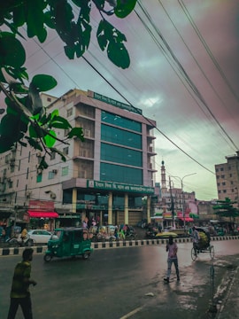 A bustling urban street scene featuring a multi-story building with a glass facade and shop signs. The road in front showcases a few pedestrians, a moving autorickshaw, and a person pushing a cart. Overhead, numerous power lines crisscross the cloudy sky, and a few green leaves frame the top corner of the image.