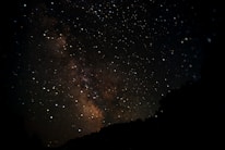 A star-filled night sky with the Milky Way galaxy prominently visible. Stars of varying brightness are scattered across the dark expanse, with the galaxy's dense cluster of stars and dust creating a hazy, luminous band. Silhouettes of trees are faintly visible at the bottom.