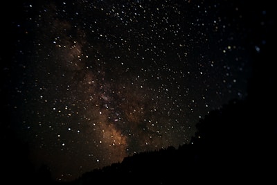 A star-filled night sky with the Milky Way galaxy prominently visible. Stars of varying brightness are scattered across the dark expanse, with the galaxy's dense cluster of stars and dust creating a hazy, luminous band. Silhouettes of trees are faintly visible at the bottom.