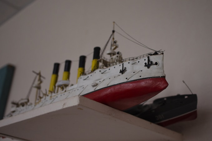 A detailed model of a classic ocean liner showcased on a shelf, featuring a white hull with red bottom paint and multiple yellow and black smokestacks. The model includes fine details such as railings and lifeboats, emphasizing the craftsmanship involved in its creation.