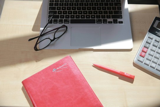 A close-up view of a workspace featuring a laptop, a pair of eyeglasses resting on the laptop's keyboard, a red notebook, and a red pen placed on a light-colored wooden desk. Additionally, there is a large calculator positioned to the right of the image.