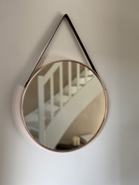 A round mirror with a thin frame is hanging on a wall, reflecting part of a white staircase in its surface. The mirror's strap appears to be leather, adding a touch of elegance to this minimalist setting.