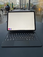 A tablet with a keyboard attachment is placed on a table in a cafe-like setting. The screen of the tablet is on, displaying a drawing or note-taking application with a blank canvas. The interior has modern, industrial design elements with large windows, allowing natural light to fill the space. Several tables and chairs are visible, along with blurred figures presumably engaged in conversation or using electronic devices.