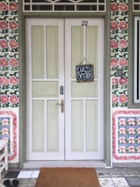 A double wooden door with a pastel green hue and the number 22 above. A small chalkboard sign hangs in the center, saying 'Class Session'. The surrounding wall features a floral-patterned wallpaper with pink and green colors. On the floor, there is a brown doormat and black shoes to the side.