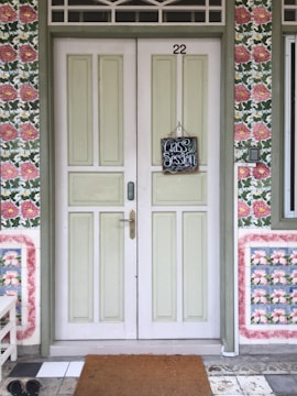A double wooden door with a pastel green hue and the number 22 above. A small chalkboard sign hangs in the center, saying 'Class Session'. The surrounding wall features a floral-patterned wallpaper with pink and green colors. On the floor, there is a brown doormat and black shoes to the side.