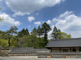 Traditional Korean architecture with a tiled roof and intricate designs is nestled among lush green trees. The sky is clear with scattered white clouds, suggesting a peaceful and serene setting.