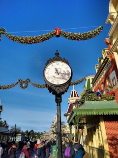A vintage-style street clock with Roman numerals is prominently displayed with festive holiday garlands and decorations. The backdrop includes colorful buildings with ornate architecture, and many people are walking in a lively pedestrian street. In the distance, a castle-like structure is visible under a clear blue sky.