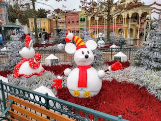 In a decorated park-like setting, a snow sculpture of a cartoonish mouse wearing a yellow and red striped hat and a red scarf stands amidst snowy foliage and bright red plants. Other sculptures resembling cartoon characters are present, alongside festive decorations such as garlands and banners adorning a colorful building facade in the background.
