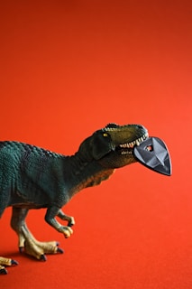 A toy dinosaur with a green textured body is depicted against a reddish-orange background. It holds a black guitar pick in its open mouth, creating a playful and imaginative scene.