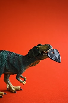A toy dinosaur with a green textured body is depicted against a reddish-orange background. It holds a black guitar pick in its open mouth, creating a playful and imaginative scene.