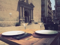 A wooden table is set outdoors with two round white plates, cutlery wrapped in napkins, and empty wine glasses. The table is located in a plaza next to an old stone building with an arched doorway. The building is adorned with graffiti, and there's a sign titled 'La Piazza' on a black board. Surrounding the area are multiple-story buildings with balconies.